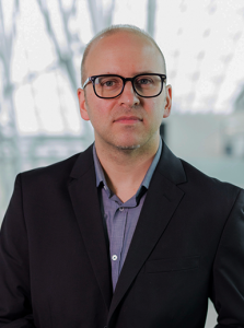 Man with black rimmed glasses, dark blue dress shirt, dark blazer, slightly smiling at the camera.